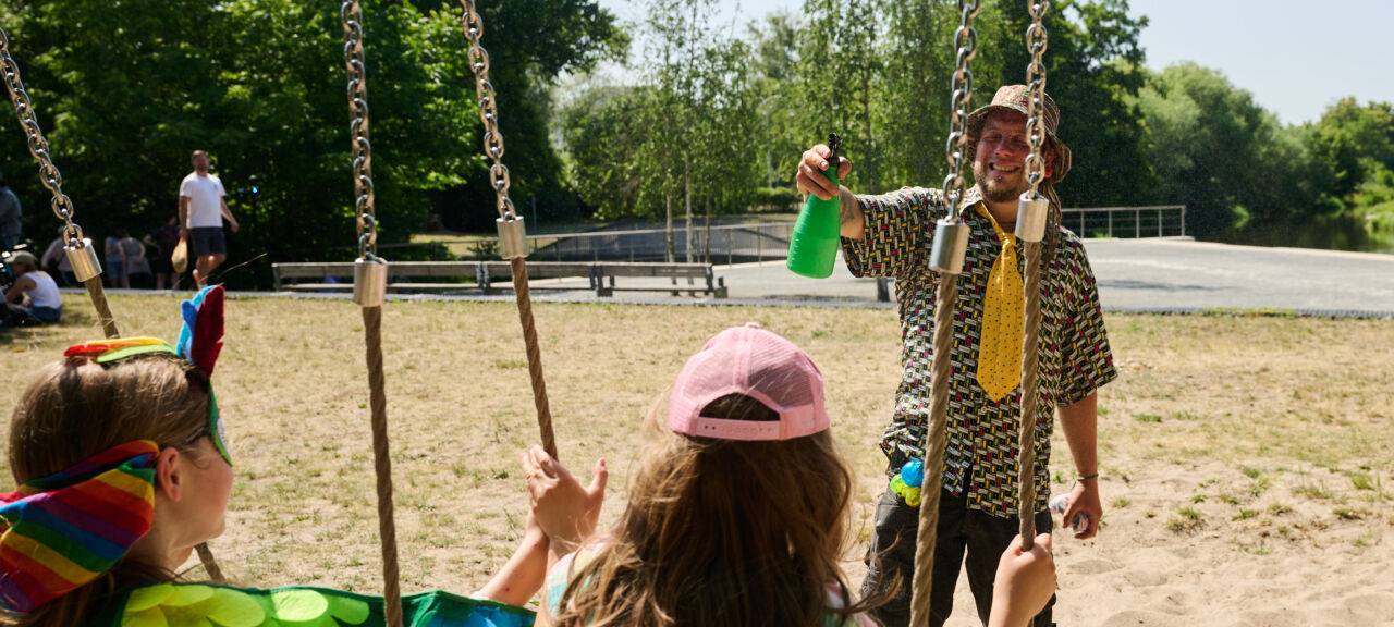 Ein Mann bespritzt seine Kinder mit Wasser, während diese auf Schaukeln sitzen. Zum Sommeranfang herrschen hochsommerliche Tempertauren.